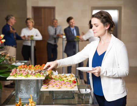 Young businesswoman serving herself with canapes in buffet of restaurant. Business people eating and talking in background